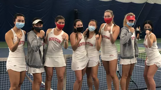 Eight members of the Boston University women's tennis team do a fist pump together in front of a tennis court net after earning a 7-0 win against Rhode Island.