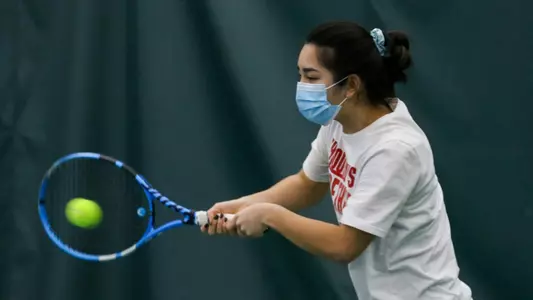 Women's Tennis player wearing a blue mask and white t-shirt hits a double handed backhand.