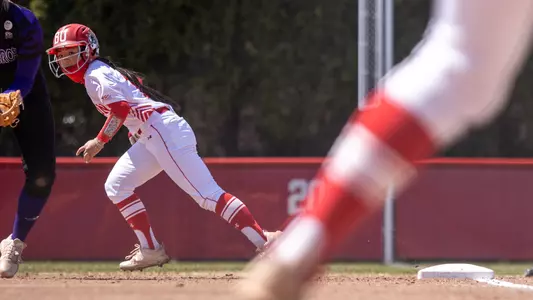 Camera is pointing from behind home plate at Jen Horita, who takes a lead at first as a pitch is delivered home.
