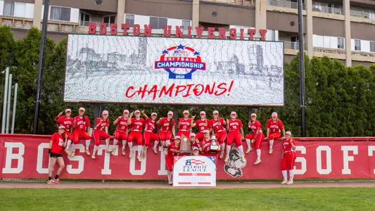 The Boston University softball players jump on the fence with the 2021 Patriot League championship trophy and banner for a photo in front of the video board declaring BU champions.