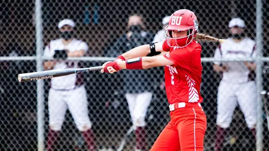 Emily Gant crushes a ball batting left handed at the plate as Lafayette players look on from their dugout protected by a chained link fence.