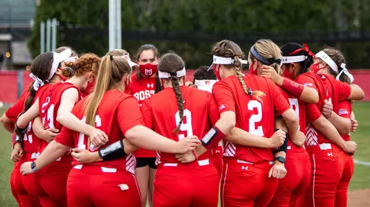 The 2021 BU softball team gathers in a large circular huddle with arms around each other.