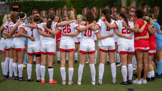 The women's soccer team in a huddle with BU ivy in the background