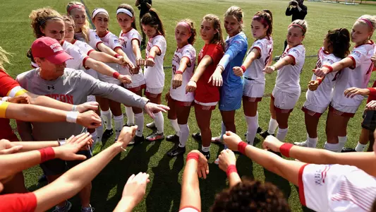 Boston University women's soccer team stretches out arms into the center of a circular huddle.