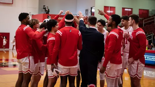 The men's basketball team gathers in a circle near the middle of the court for a team huddle.
