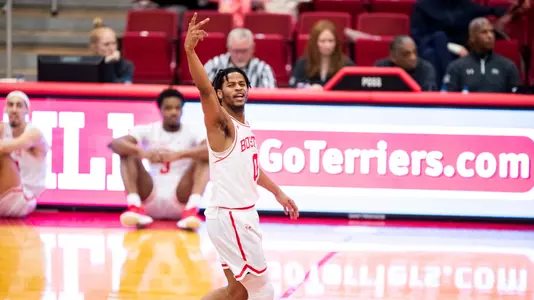 Ethan Brittain-Watts celebrates hitting a 3-pointer by raising his fingers in the air toward the fans in the stands.