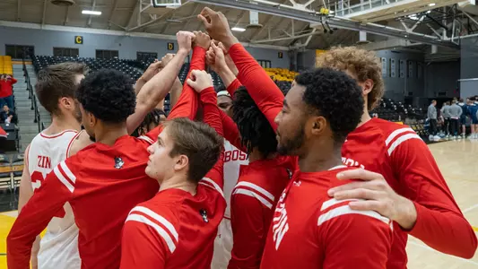 Men's Basketball Team Huddles with arms outstretched on the court.