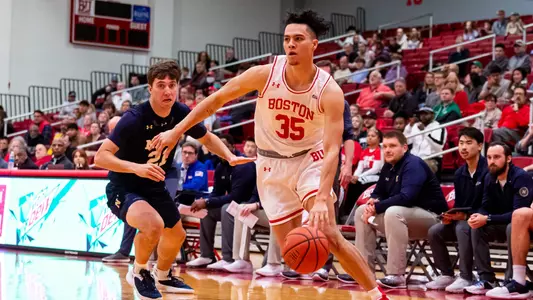 Fletcher Tynen dribbles toward the hoop in front of Navy bench with a defender to his side.