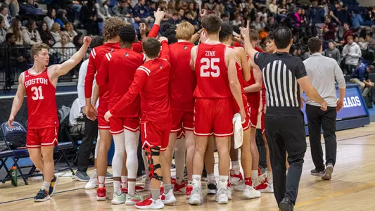 Men's Basketball team huddles with arms outstretched into middle in front of bench.