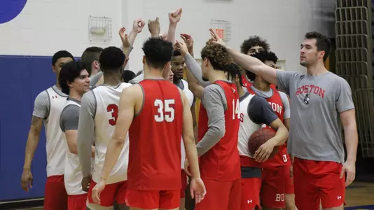2022 Boston University men's basketball team huddles with outstretched arms raised in the air during a practice.