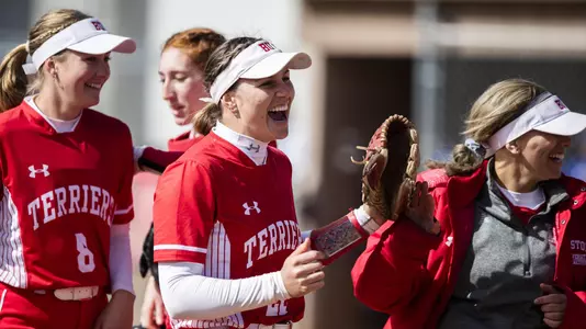 Several BU softball players walk toward the dugout smiling and cheering after ending the top of an inning with the third out.
