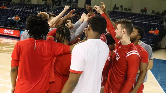 Men's Basketball team huddles with arms outstretched high up next to the basket during warm-ups at Bucknell.