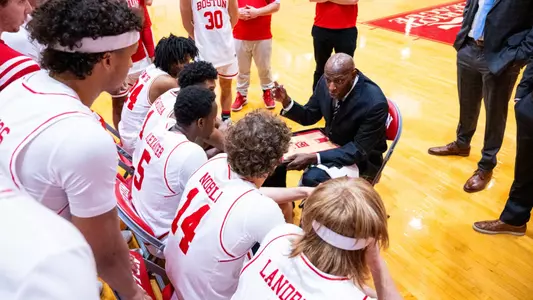 Head coach Joe Jones is sitting down on a chair in a team huddle drawing up a play while giving instruction.