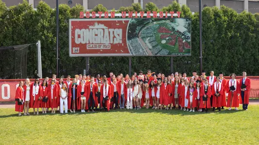 The 2023 Boston University Student-Athlete Class of 2023 gather in front of softball's video board for a group photo in cap and gown.