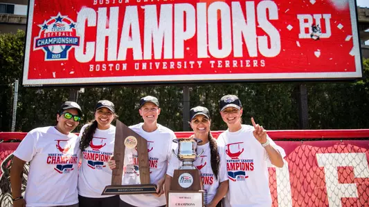 Boston University softball coaching staff pose for a group photo in front of the video board with the two Patriot League championship trophies.