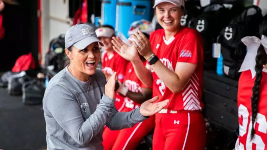 BU softball head coach Ashley Waters laughs in the dugout with multiple players while clapping her hands.