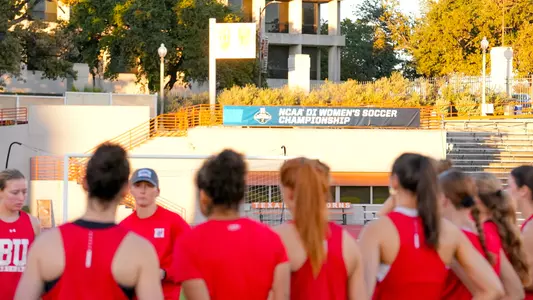 Photo of a BU women's soccer team huddle at the Mike A. Myers Stadium in Austin, Texas, with the NCAA Championship banner in the background.