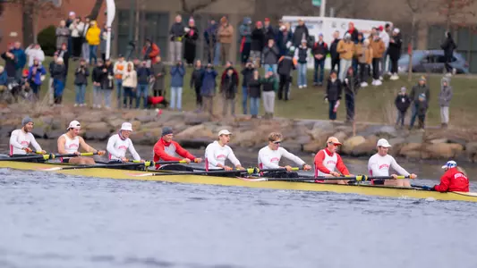 Men's Rowing eights boat rows down the Charles River with fans looking on from the Cambridge side.