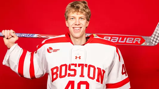 Mikhail Yegorov posing ands smiling in his white BU jersey while holding his stick behind him