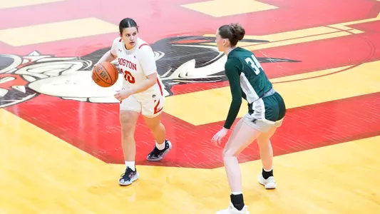Photo of women's basketball senior Alex Giannaros dribbling the ball with a Loyola Maryland defender facing her.