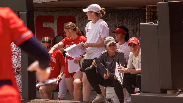 Photo shows several players and the coaching staff in the dugout with two coaches sitting down looking toward home plate with the team out on the field
