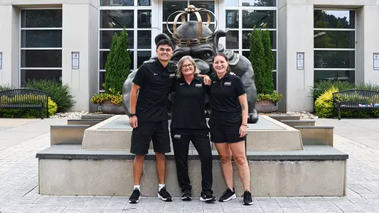 Ethan Larsen, Christy Morgan, and Morgan DeMann pose together for coaches' photo in front of the Duke Dog statue in front of the Robert & Frances Plecker Athletic Performance Center