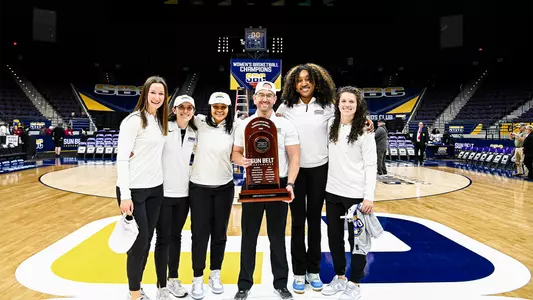 Anastasiia Zakharova, Alex Danas, Nicole Razor, Sean O'Regan, Kayla Cooper Williams, and Alex Tomlinson posing with the Sun Belt Tournament trophy on the court at the Pensacola Bay Center.