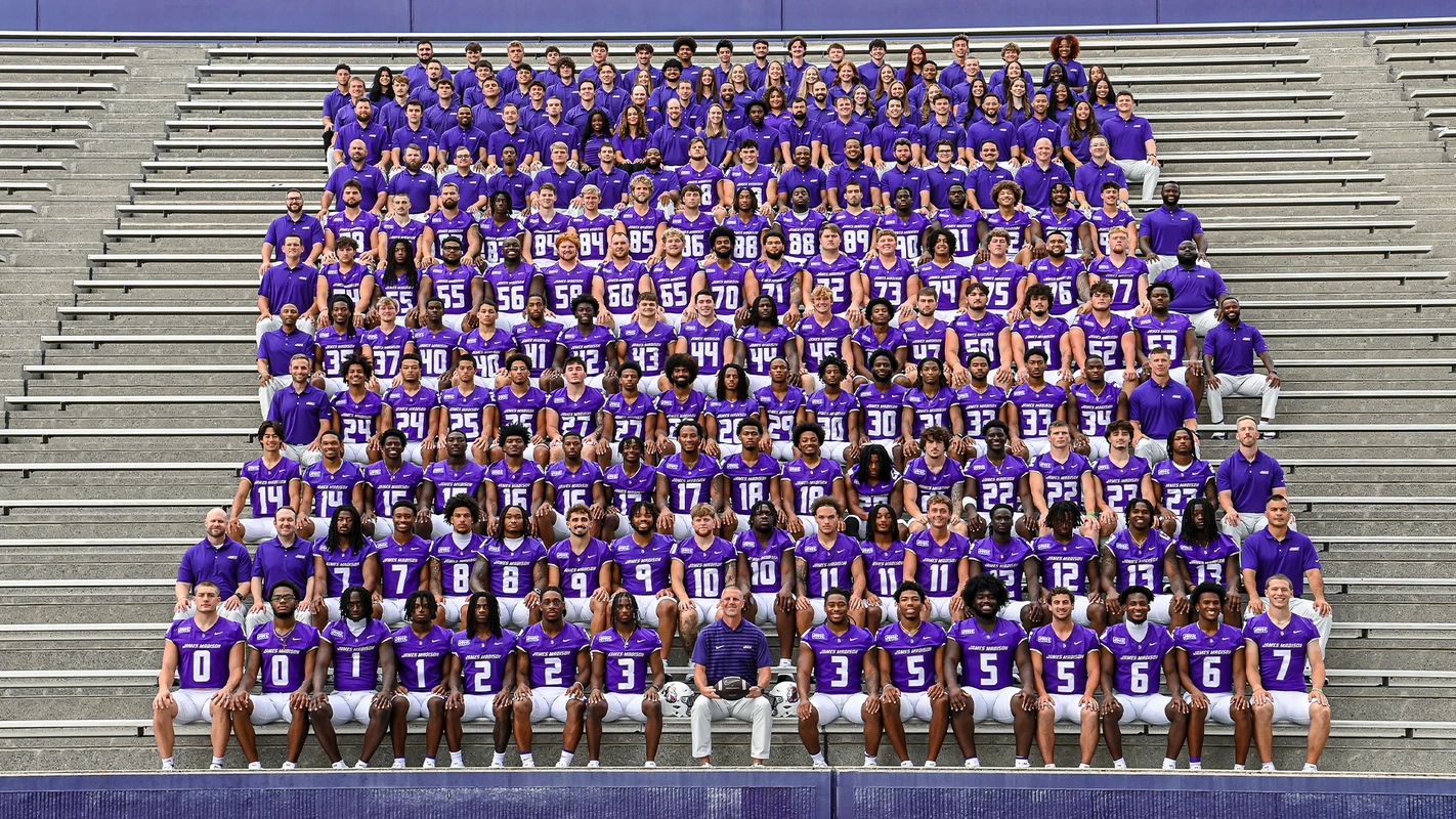 JMU football players and coaches in uniform sitting in the stands at Bridgeforth Stadium for the team photo.