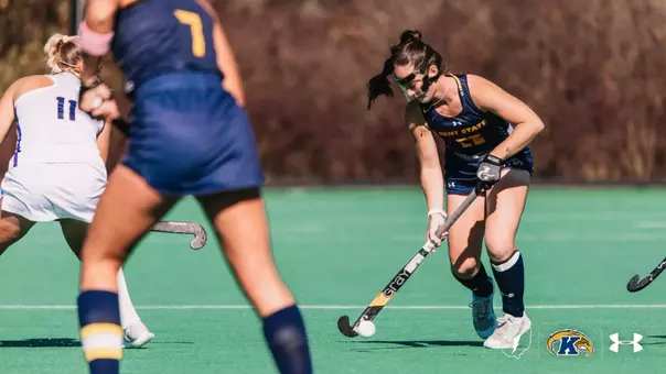 Kent State field hockey player No. 26, wearing a mask and gloves, dribbles the ball down the field while pressured by two JMU defenders during a match on the turf.