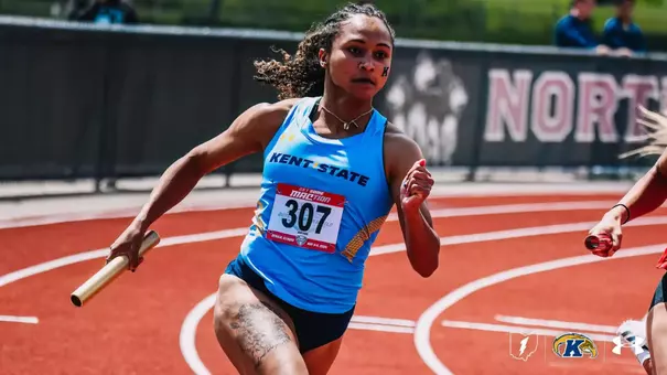 Kent State University track and field athlete Amryne Chilton runs a relay leg at the 2025 MAC Outdoor Track and Field Championships in DeKalb, Illinois, baton in hand and mid-stride on the red track surface. Chilton wears a light blue Kent State uniform and bib number 307. The Kent State Athletics logo and Under Armour wordmark appear in the bottom right corner.