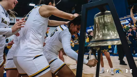 Kent State women's basketball players celebrate a win by ringing the victory bell courtside, teammates cheering and reaching in as one player pulls the rope, with cheerleaders and fans visible in the background.