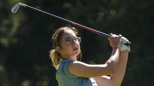 Kent State women’s golfer Petra Babicová follows through on a swing during competition, wearing a blue sleeveless top and dark shorts, with trees blurred in the background.