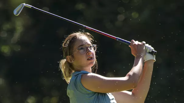 Kent State women’s golfer Petra Babicová follows through on a swing during competition, wearing a blue sleeveless top and dark shorts, with trees blurred in the background.