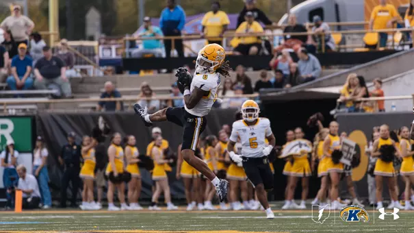 An action shot of a Kent State football player wearing a white jersey and yellow helmet, leaping high into the air to catch a pass during a game, with cheerleaders and spectators in the background.