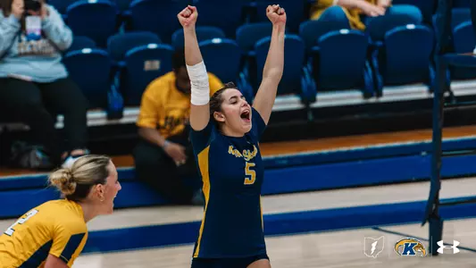 Kent State volleyball player #5 in a navy and gold jersey raises both fists overhead and yells in celebration, with a white wrap on her forearm. A teammate in a gold jersey leans in at the lower left, also cheering, while fans sit in the blue bleachers behind them.