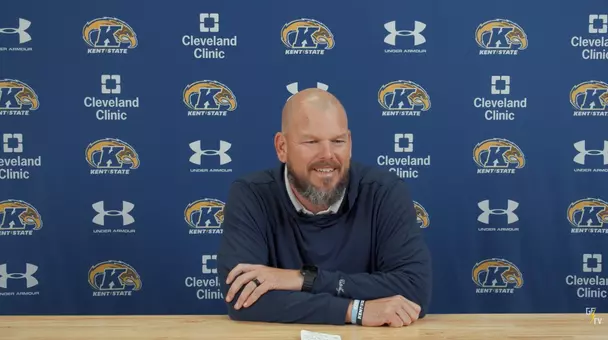 A bald man with a beard smiling while seated at a wooden desk in front of a blue Kent State press backdrop featuring repeating logos for Kent State, Under Armour, and Cleveland Clinic.
