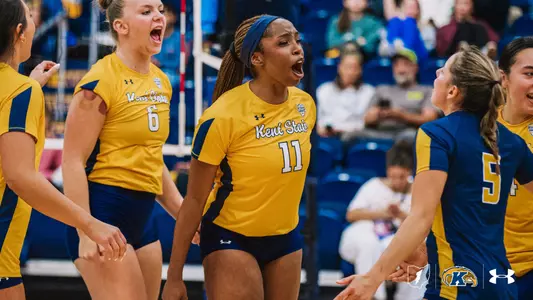 Kent State volleyball player #11 yells in celebration at the center of the huddle, with teammate #6 shouting beside her and #54 in a navy jersey reaching in from the right. The players wear gold Kent State jerseys, and spectators fill the stands behind them.