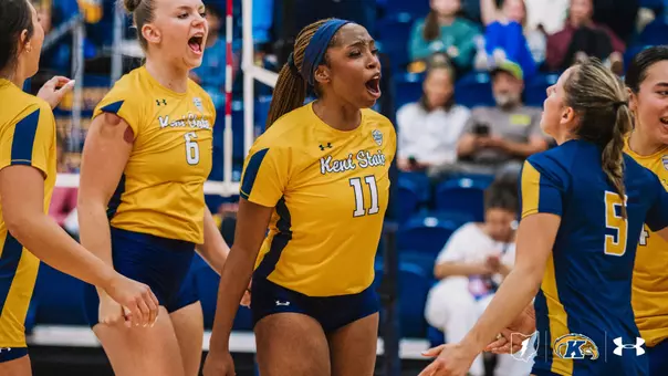 Kent State volleyball player #11 yells in celebration at the center of the huddle, with teammate #6 shouting beside her and #54 in a navy jersey reaching in from the right. The players wear gold Kent State jerseys, and spectators fill the stands behind them.