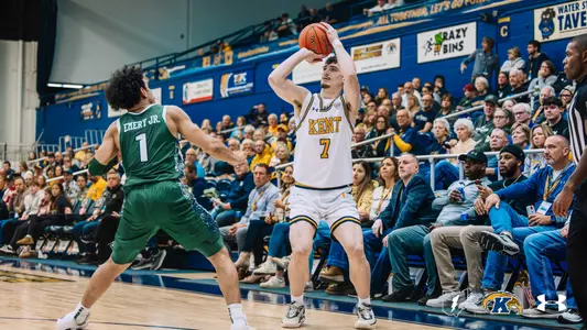 Kent State player #7, in a white home jersey with gold and navy trim, rises into a three-point shooting motion with the ball overhead while Emery Jr. (#1) of the opposing team, in a green jersey, defends. Fans fill the stands behind them.