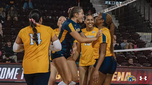Kent State volleyball players in gold and navy uniforms celebrate together on the court at Central Michigan, smiling and cheering as they gather near the net. A player in a gold #14 jersey stands with her back to the camera in the foreground, while a teammate in a navy jersey leans in with her hand on another player’s shoulder.