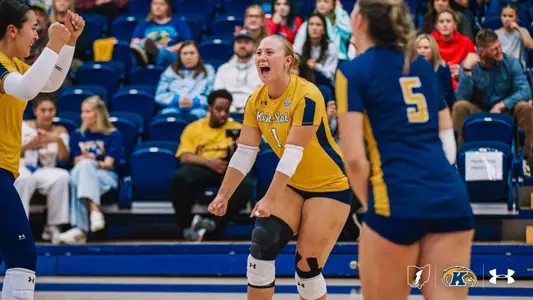 Kent State volleyball player in a gold No. 1 jersey crouches forward with clenched fists and shouts in celebration. A teammate in gold smiles with raised fists on the left, and a teammate in a navy No. 5 jersey stands in the foreground with her back to the camera. Spectators watch from the blue stands behind them.