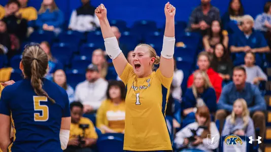 Kent State volleyball player in a gold No. 1 jersey raises both fists above her head and yells in celebration, while a teammate in a navy No. 5 jersey stands in the foreground with her back to the camera. Fans fill the blue bleachers behind them.