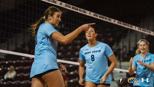 Kent State volleyball player in a light blue jersey points emphatically while smiling and talking during a match at Central Michigan, as two teammates in matching jerseys, including #8, celebrate behind her.
