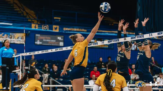 Kent State volleyball’s No. 6, in a gold jersey, leaps and reaches high with her right arm to tap the ball over the net as two Akron players in navy jerseys, Nos. 16 and 14, jump with hands raised to block. Two Kent State teammates, Nos. 12 and 4, watch from the foreground.