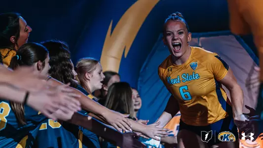 Kent State volleyball’s No. 6, in a gold jersey, runs out of an inflatable tunnel during player introductions, shouting with excitement as teammates lined up along the side reach out for high fives.