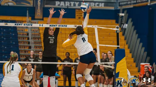 Kent State volleyball’s No. 8, in a white jersey with gold sleeves, leaps and swings her right arm back to hit the ball as an opposing blocker in a black jersey jumps with hands raised at the net. Teammates Nos. 7 and 5 look on from behind the net.