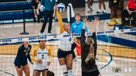 Kent State volleyball’s No. 8, in a white jersey with gold sleeves, leaps toward the net to attack the ball as an opposing blocker in a dark jersey, No. 23, jumps with arms raised. A Kent State teammate, No. 1, watches in the foreground, and the opposing bench is visible in the background.