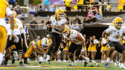 Kent State quarterback scrambling with the football during a game while teammates block around him.
