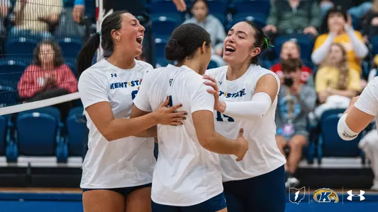 Three Kent State volleyball players in white jerseys celebrate together near the net, smiling and cheering as two of them embrace a teammate whose back faces the camera, with fans seated in the blue arena stands behind them.