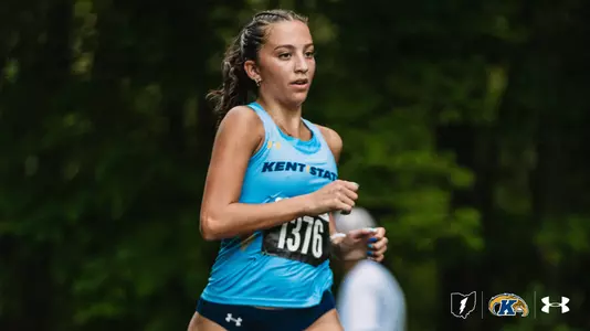 Kent State women's cross country runner in a light blue singlet with bib number 1376 runs along a shaded, wooded course, her braided ponytail swinging behind her.